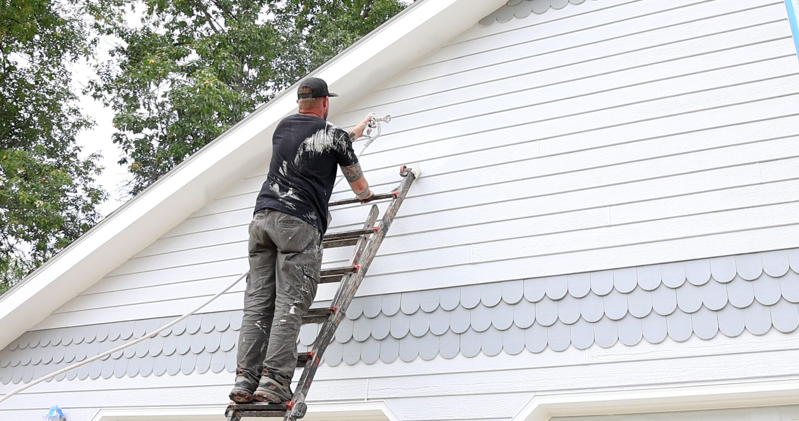 Painter with Graco Sprayer sprays grey paint on white home during summer.