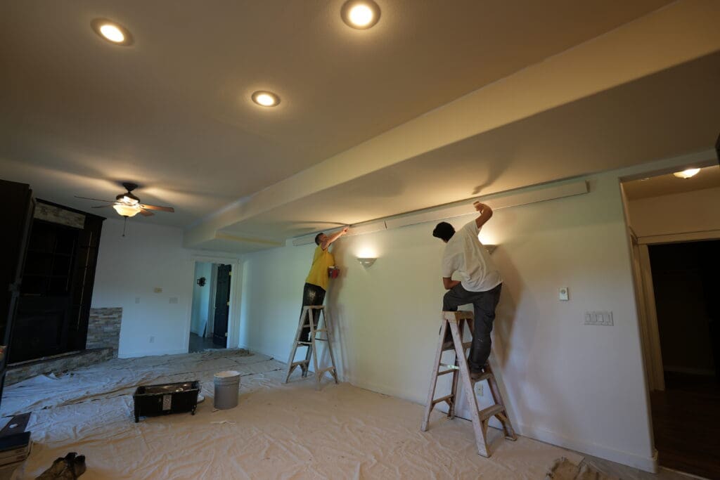 Two painters from Black Mountain Painting work on a basement interior in a home in Belle Fourche, South Dakota. Both are standing on ladders, painting the ceiling and upper walls, with drop cloths covering the floor for protection.
