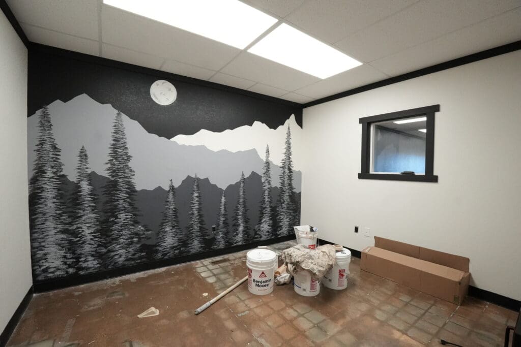 A custom wall mural painted by Black Mountain Painting for Black Mountain Cleaners in Spearfish, SD, featuring a grayscale mountain and pine tree landscape with a moon above, shown inside a room with painting supplies on the floor.