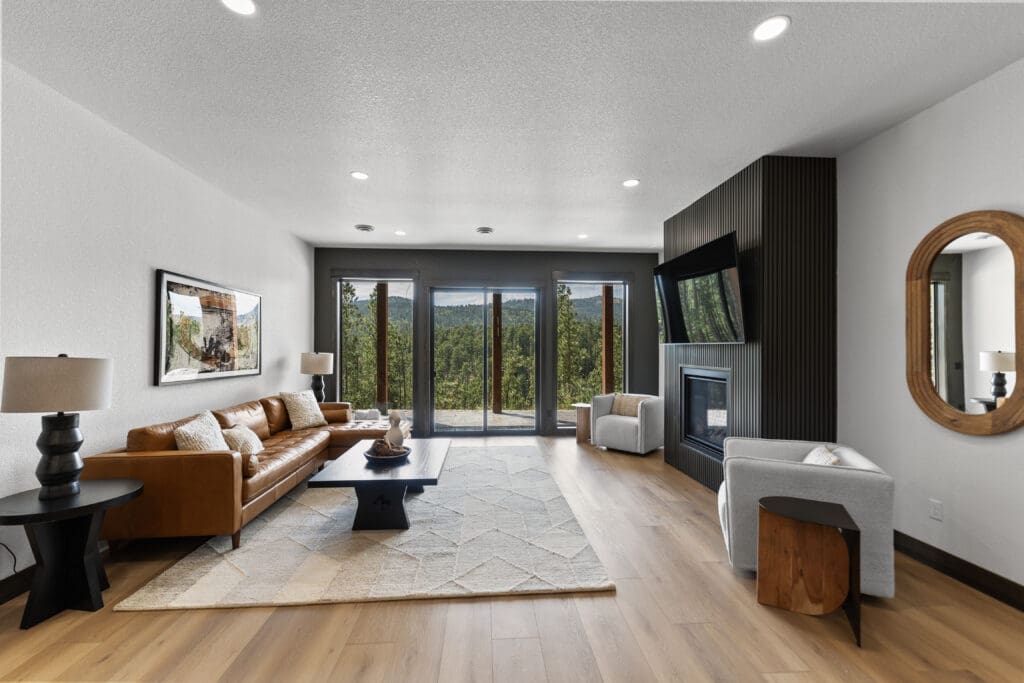 Living room with white walls, a charcoal window wall, and a dark slatted fireplace surround opening to a forest view in a Powder House Pass home near Lead, SD.